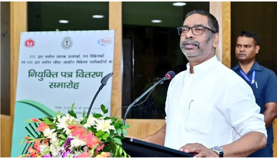 A government official speaking at an appointment letter distribution ceremony, standing at a podium with floral decoration.